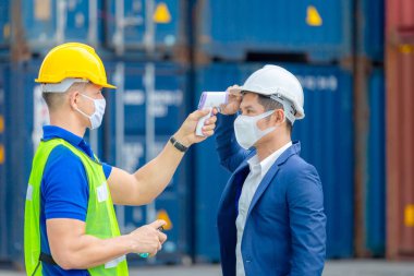 Kargo konteynerlerindeki Coronavirus (COVID-19) taraması ve korunması için ateşi dijital termometreyle kontrol eden fabrika işçisi