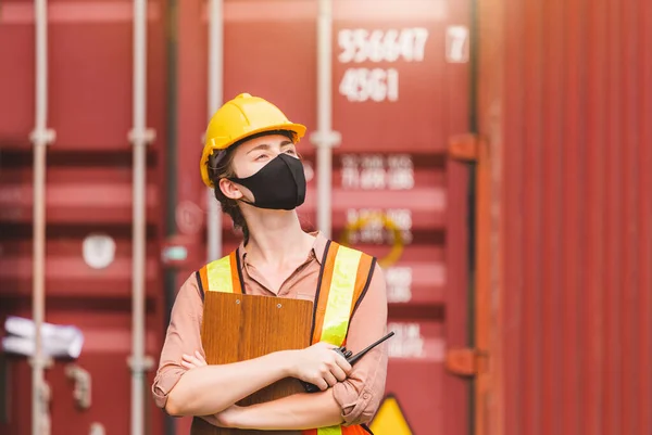 Engineer in hardhat and safety vest holding clipboard checklist and two-way radio, Woman wearing protection face mask against coronavirus at containers cargo