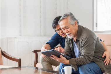 Yaşlılar ve teknoloji konsepti, Asyalı kıdemli baba ve yetişkin oğul oturma odasında tablet akıllı telefon kullanıyor.