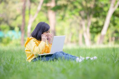 Neşeli küçük kız çimlerin üzerinde oturuyor, parkta dizüstü bilgisayarla çalışıyor, açık havada öğreniyor.