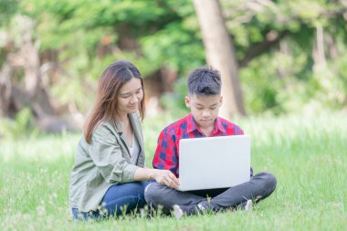 Neşeli anne ve oğul çimlerin üzerinde oturur, parkta dizüstü bilgisayarla çalışır, açık havada öğrenim kavramı geliştirir.