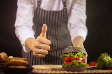 Şef parmaklarını gösterip elinde bir hamburger tutuyor, ev yapımı fırın konsepti.