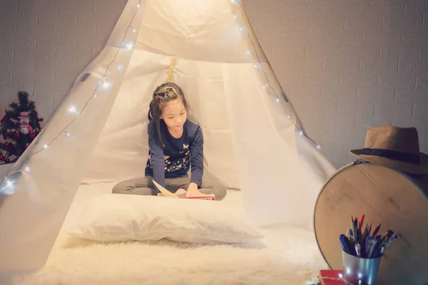 Little girl reading a book in tent, Happy kid playing at home, Funny lovely kid having fun in children room.