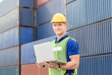 Kask ve güvenlik yeleği giyen işçi dizüstü bilgisayar tutuyor Foreman konteynır kutusunu kargodan yüklüyor.