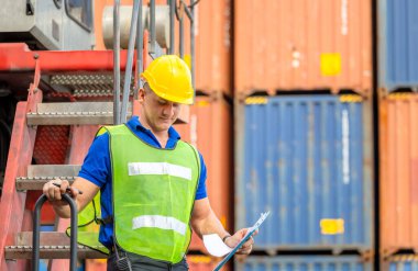 Kask ve güvenlik yeleği giymiş işçiler konteyner stackers üzerinde duruyor. Yükleme konteynerlerini kargo kutusundan kontrol ediyorlar.