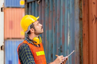 Kara şapkalı fabrika işçisi elinde pano listesi ve kargo kutusundan konteynır kutusunu kontrol ediyor.