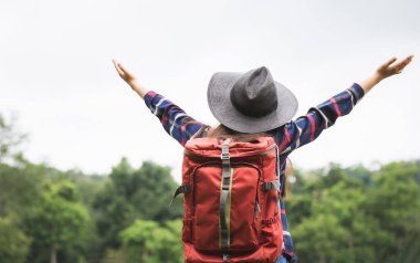 Sırt çantası ve şapkası olan genç bir kadının arka görüntüsü Gezgin, kollarını kaldırıp güzel bir doğanın tadını çıkarıyor.