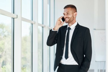 Gülümseyen yakışıklı işadamı, açık alanda panoramik pencereli modern ofiste telefonla konuşuyor.