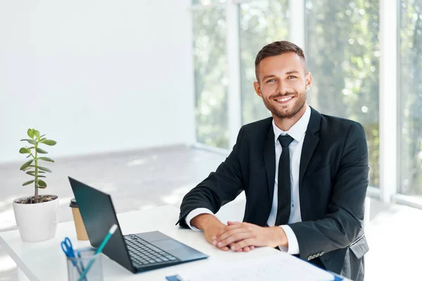 Snygg leende affärsman i elegant kostym sitter vid sitt skrivbord i en ljus modernt kontor — Stockfoto