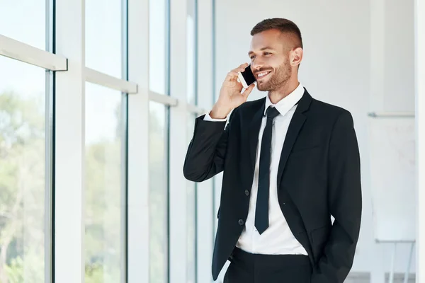 Sonriente hombre de negocios guapo hablando por teléfono en espacio abierto oficina moderna con ventanas panorámicas de fondo — Foto de Stock