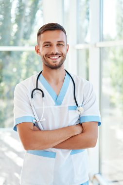 Tıbbi üniformalı, steteskoplu, kendinden emin gülümseyen bir erkek doktorun portresi.