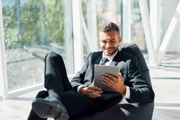 Zarif süitte gülümseyen başarılı iş adamı yaratıcı ofisteki fasulye çuvalının üzerinde tablet kullanıyor. — Stok fotoğraf