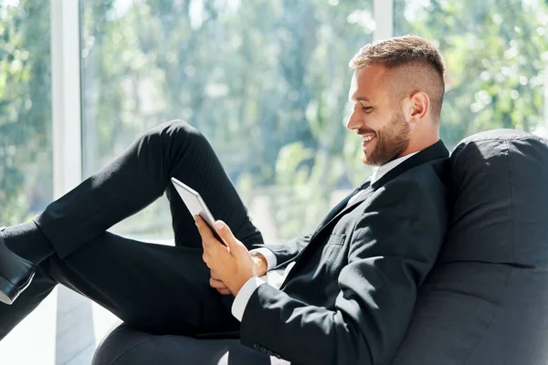Sonriente exitoso hombre de negocios en elegante suite usando tableta sentada en una silla de bolsa de frijol en una oficina creativa — Foto de Stock