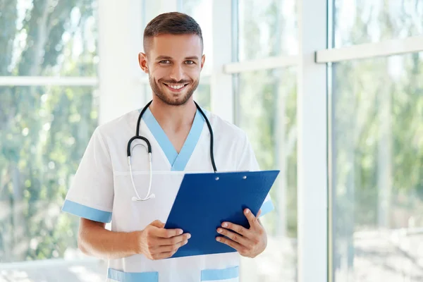 Portræt af selvsikker mandlig læge med stetoskop og udklipsholder i medicinsk uniform - Stock-foto