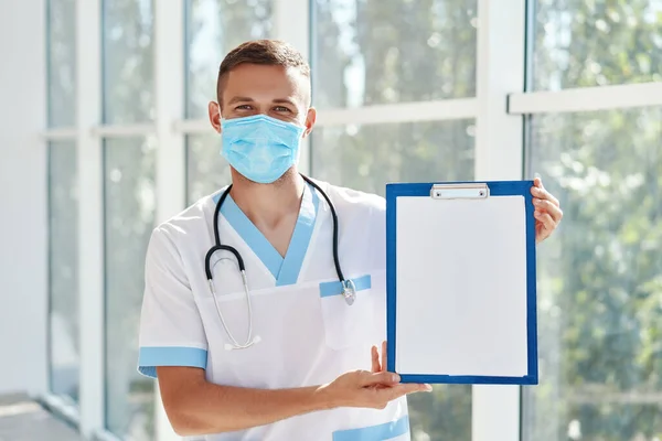 Selvsikker mandlig læge iført medicinsk maske viser udklipsholderen med diagnose i hospitalets baggrund. - Stock-foto