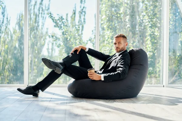 Geschäftsmann entspannt sich auf einem Sitzsack-Stuhl mit einer Tasse Kaffee vor dem Hintergrund von Panoramafenstern im modernen Büro — Stockfoto