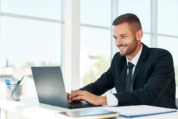 Przystojny uśmiechnięty biznesmen w eleganckim garniturze pracujący na laptopie w jasnym nowoczesnym biurze — Zdjęcie stockowe