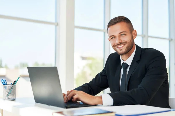 Hezký usmívající se podnikatel v elegantním obleku pracuje na notebooku v jasné moderní kanceláři — Stock fotografie