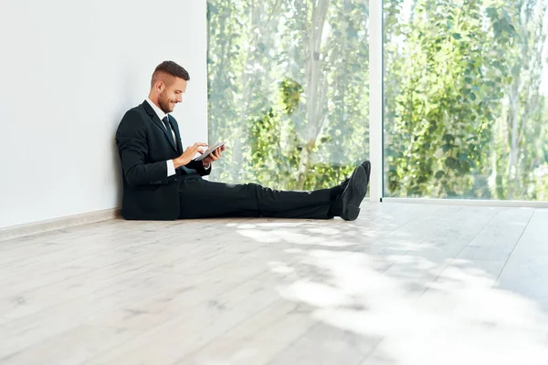 Jonge glimlachende zakenman zit op de vloer in zijn kantoor en kijkt naar de smartphone — Stockfoto