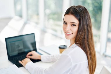 Güzel bir iş kadını masasında çalışırken kameraya bakıyor ve gülümsüyor.