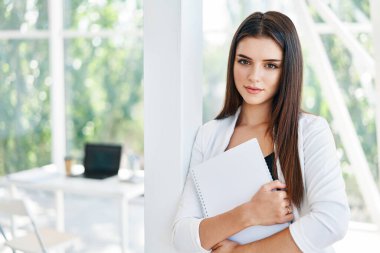 Belgeleri tutan ve modern yaratıcı ofiste kameraya bakan kendine güvenen bir iş kadını.