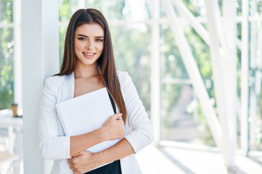 Belgeleri tutan ve modern yaratıcı ofiste kameraya bakan kendine güvenen bir iş kadını.