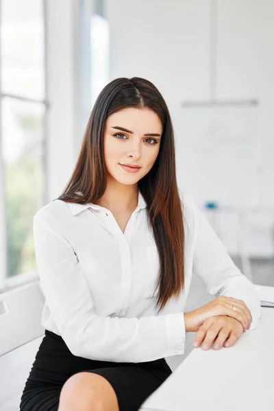 Portrait of smiling pretty young businesswoman sitting on workplace at the table in modern office and looking at camera — Stock Photo, Image