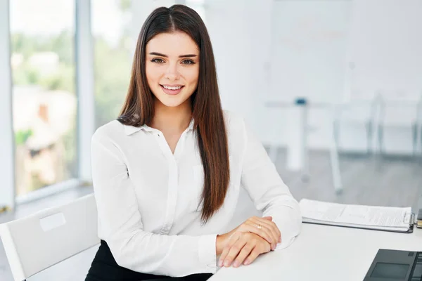 Porträt einer lächelnden hübschen jungen Geschäftsfrau, die am Arbeitsplatz am Tisch im modernen Büro sitzt und in die Kamera blickt — Stockfoto