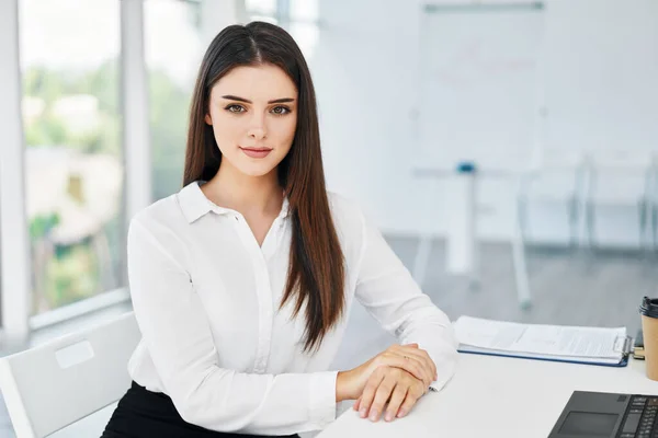 Retrato de sorrir muito jovem empresária sentada no local de trabalho à mesa no escritório moderno e olhando para a câmera — Fotografia de Stock