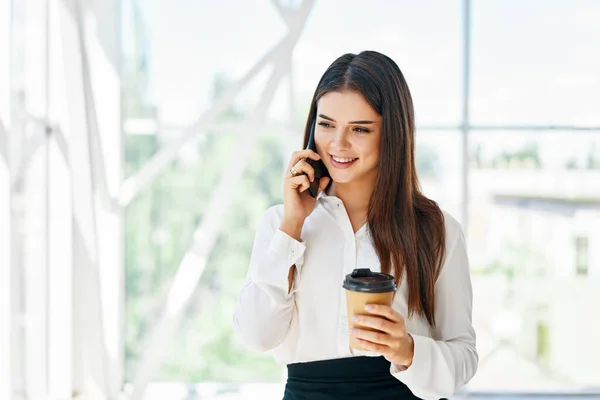 Smilende vakker forretningskvinne som snakker i telefonen med kaffekopp på det moderne kontoret. stockbilde
