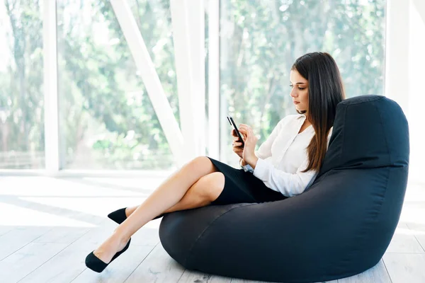 Wanita cantik yang percaya diri menggunakan telepon genggam menjawab pesan-pesan penting yang duduk di kursi kantong kacang Stok Gambar Bebas Royalti