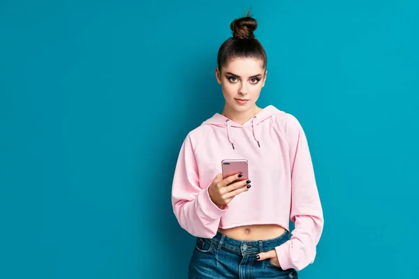 Bastante chica hipster millennial sosteniendo el teléfono móvil y mirando a la cámara en fondo de estudio azul — Foto de Stock