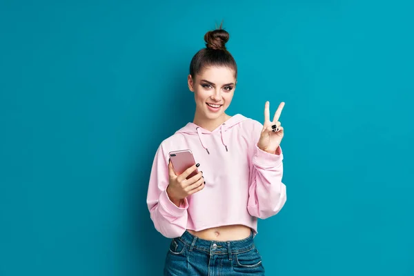 Hübsches Millennial-Hipster-Mädchen hält Handy in der Hand und macht Siegeszeichen — Stockfoto