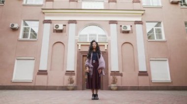 Genç, güzel Asyalı bir kadın büyük apartmanların yakınında kameraya bakıyor.