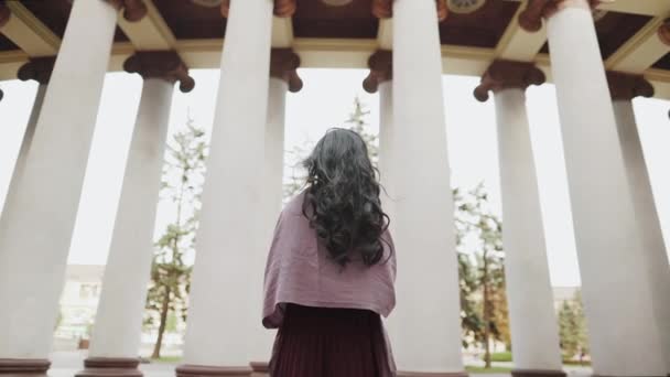 Vue arrière de jeune jolie wonan aux cheveux longs restant près des colonnes regardant la rue — Video