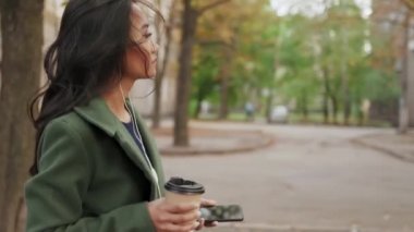 Yörüngede gülümseyen Asyalı kadının akıllı telefonuyla müzik dinlediği ve kahve içtiği bir fotoğraf var.