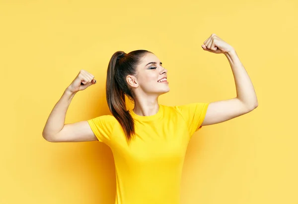 Kraftfull säker ung kvinna visar armar muskler på gul bakgrund — Stockfoto