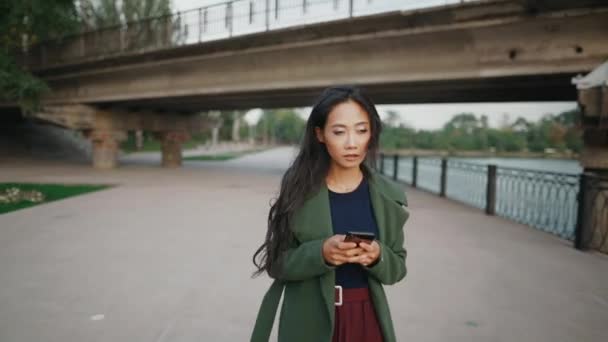 강둑을 걸어 다닐 때새로운 앱을 온라인으로 사용하여 스마트폰으로 문자 메시지를 보내는 행복 한 아시아인 — 비디오