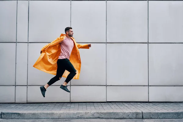 Modny młody człowiek w żółtym płaszczu przeciwdeszczowym skacze w powietrzu na ścianie ulicy tła miejskiego — Zdjęcie stockowe