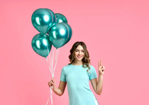 Happy pretty woman with blue balloons in hands doing victory sign on pink background — Stock Photo, Image