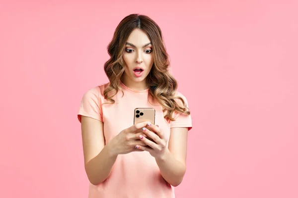 Portret van een geschokt mooi millennial hipster meisje met behulp van mobiele telefoon op roze achtergrond — Stockfoto