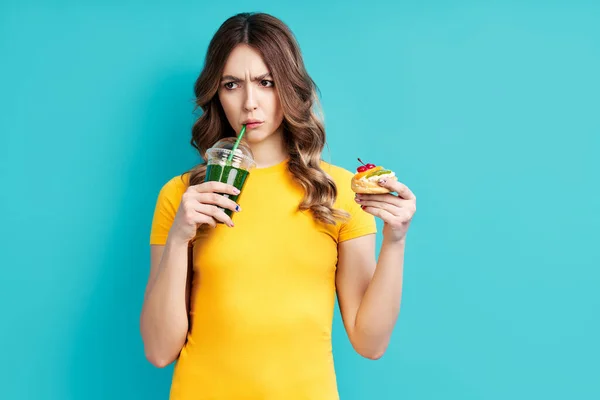 Mulher infeliz na dieta beber suco de desintoxicação segurando bolo na mão escolher alimentos saudáveis — Fotografia de Stock