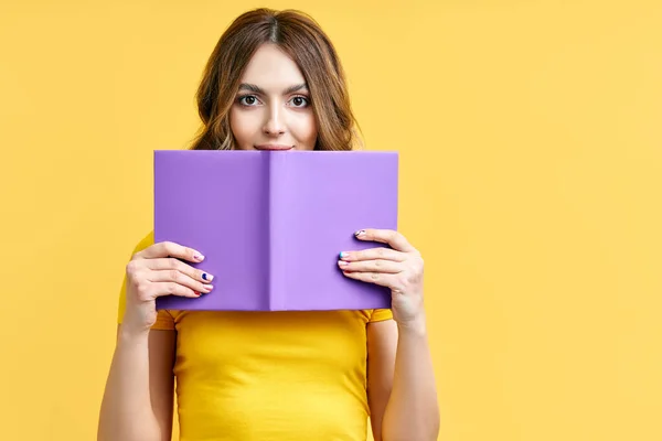 Молодая женщина прячется за открытой книгой и смотрит в камеру изолированы на желтом фоне. — стоковое фото