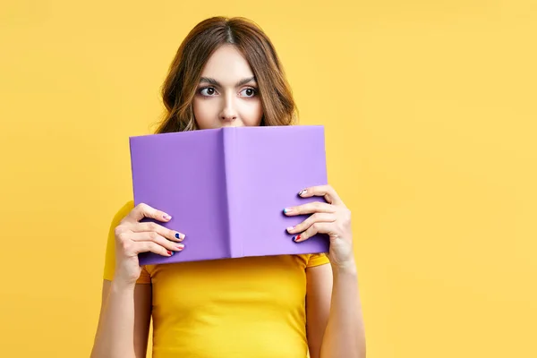 Jeune femme cachée derrière un livre ouvert et regardant la caméra isolée sur fond jaune — Photo