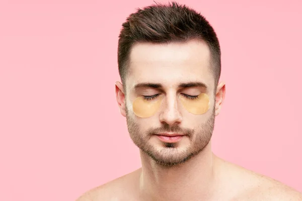 Joven hombre guapo con los ojos cerrados y parches de ojos dorados sobre fondo rosa — Foto de Stock