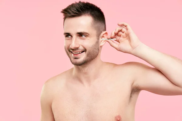 Młody uśmiechnięty mężczyzna czyści ucho wacikiem na różowym tle — Zdjęcie stockowe