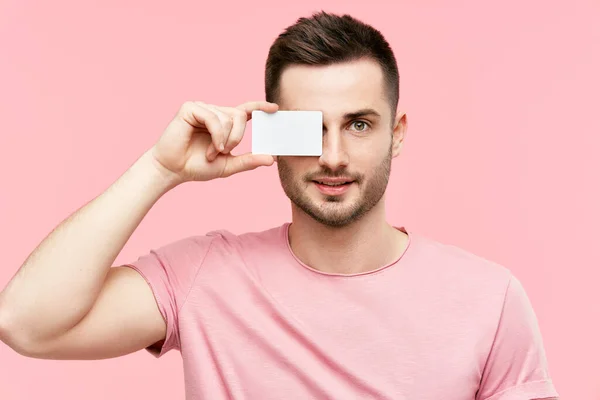 Knappe glimlachende man houdt credit card in de buurt van zijn gezicht sluiten oog over roze achtergrond — Stockfoto
