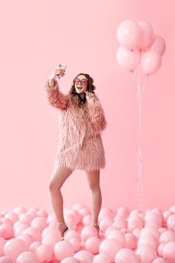 Pembe balonlar üzerinde akıllı telefondan fotoğraf çeken alımlı genç bir kadının tam boy portresi.
