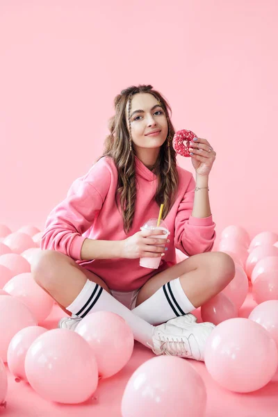 Glimlachende jonge vrouw met milkshake en donut in handen zittend op de vloer met veel roze luchtballonnen — Stockfoto