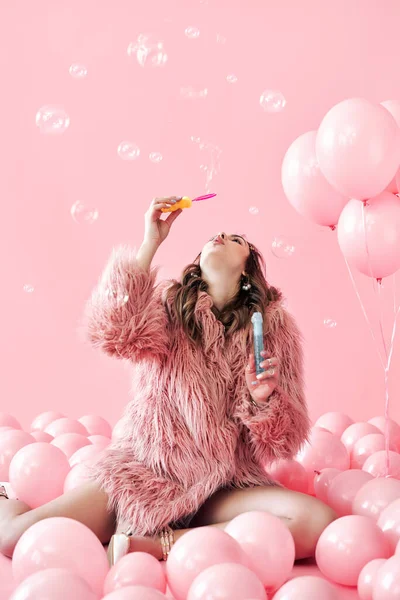 Jong mooi vrouw blazen zeepbellen op roze achtergrond met ballonnen — Stockfoto